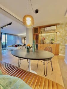 Comedor informal integrado al living, con una mesa redonda negra de base blanca acompañada por sillas metálicas y un banco tapizado en cuero color caramelo. Sobre la mesa hay dos jarrones decorativos y un bowl con frutas. Del techo cuelga una luminaria ámbar de diseño contemporáneo. Al fondo se observa una barra de madera con estantes iluminados, papel tapiz botánico en tonos dorados y un equipo de aire acondicionado. A la izquierda se ve parte del living con un sofá blanco y cortinas translúcidas que dejan