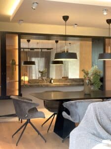 Comedor contemporáneo con mesa negra de líneas rectas y cuatro sillas tapizadas en terciopelo gris. Sobre la mesa cuelgan dos luminarias negras con interior dorado. Al fondo se ve una barra revestida en mármol con objetos decorativos y un gran espejo que amplía el espacio reflejando las cortinas del living. A la izquierda, un mueble alto con iluminación cálida interior exhibe piezas decorativas. El ambiente combina texturas suaves, tonos neutros y luz cálida, creando una atmósfera elegante y acogedora.