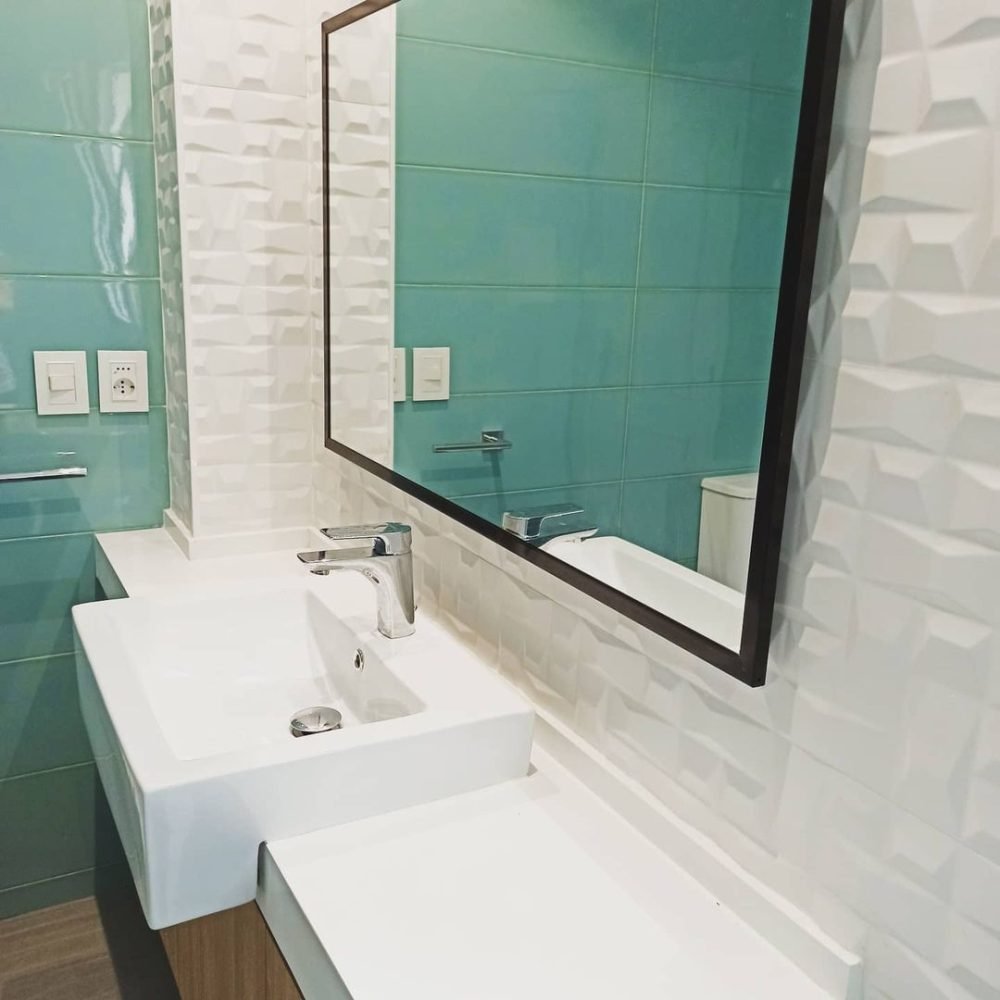 Baño contemporáneo en el barrio del Prado, Montevideo. Revestimientos en cerámica verde aguamarina, mesada de cuarzo blanco convertida en banco, mueble de roble claro, y cabina de ducha con cristal transparente y perfilería negra. Iluminación ambiental detrás del espejo y mueble flotante para crear un ambiente acogedor y moderno.