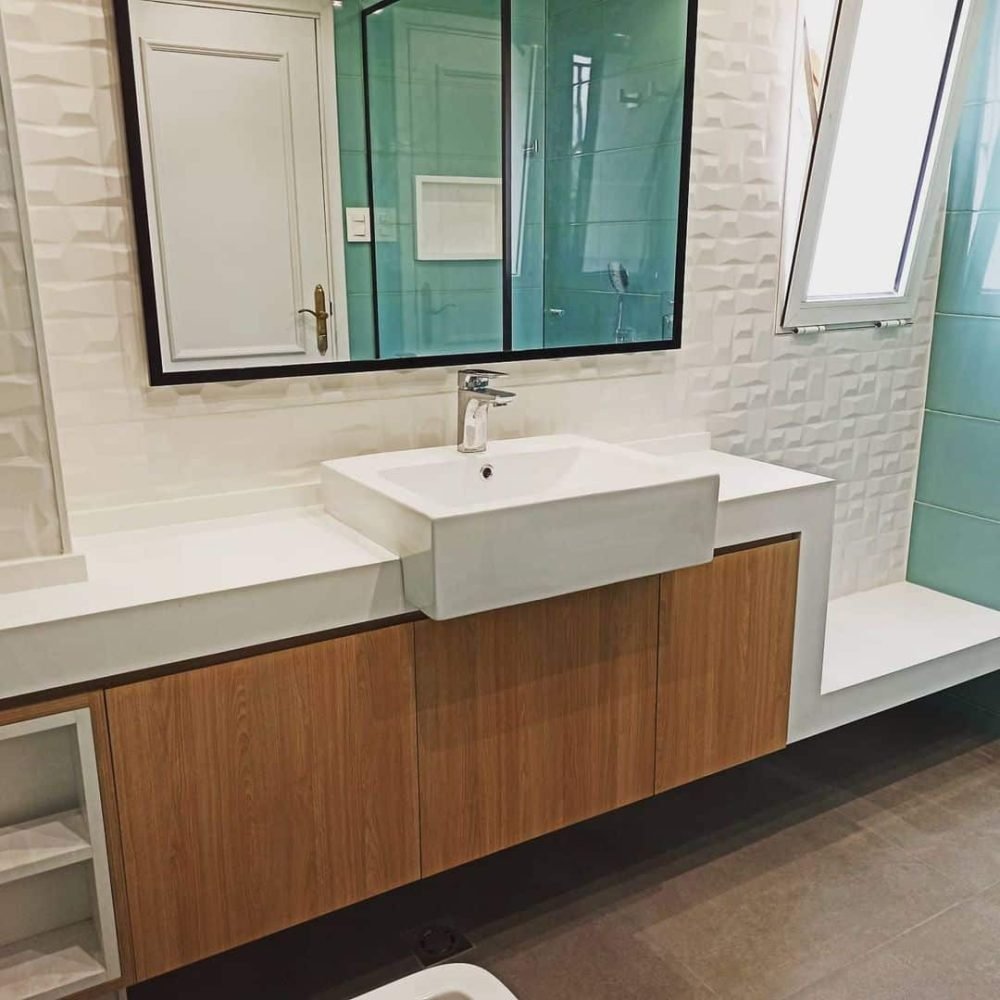 Baño contemporáneo en el barrio del Prado, Montevideo. Revestimientos en cerámica verde aguamarina, mesada de cuarzo blanco convertida en banco, mueble de roble claro, y cabina de ducha con cristal transparente y perfilería negra. Iluminación ambiental detrás del espejo y mueble flotante para crear un ambiente acogedor y moderno.