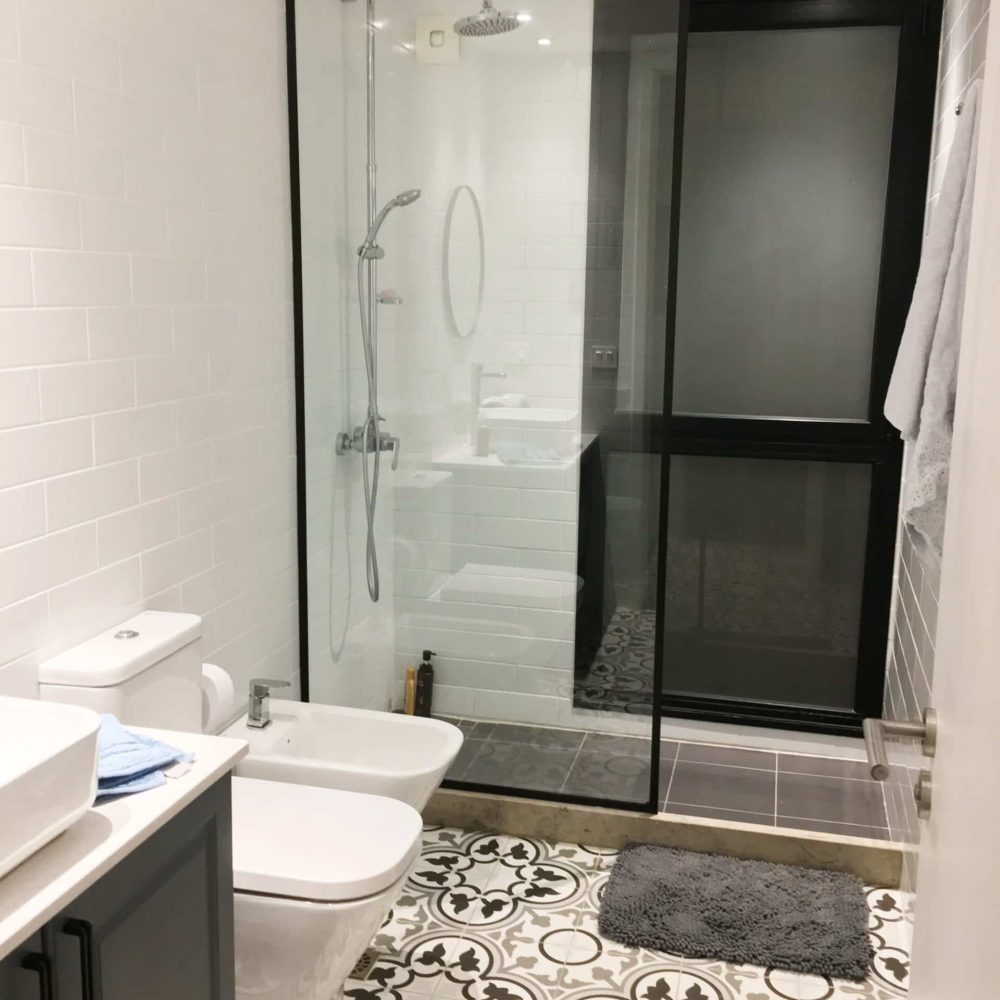 Baño contemporáneo en La Tahona, Canelones, Uruguay. Tonos grises y cálida iluminación, pisos de baldosa hidráulica. Mueble en silestone blanco y madera laqueada gris piedra. Espejo redondo con marco negro y mampara de ducha de vidrio amplio con perfilería en negro, destacando por su estilo moderno y funcionalidad.