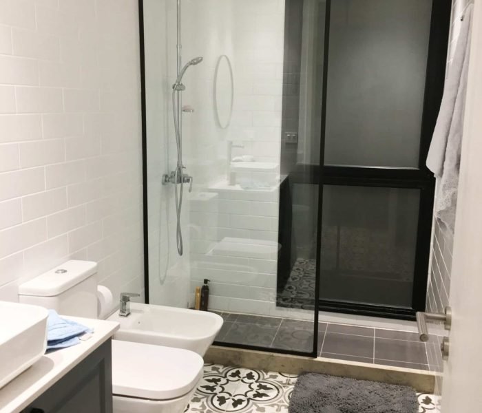 Baño contemporáneo en La Tahona, Canelones, Uruguay. Tonos grises y cálida iluminación, pisos de baldosa hidráulica. Mueble en silestone blanco y madera laqueada gris piedra. Espejo redondo con marco negro y mampara de ducha de vidrio amplio con perfilería en negro, destacando por su estilo moderno y funcionalidad.