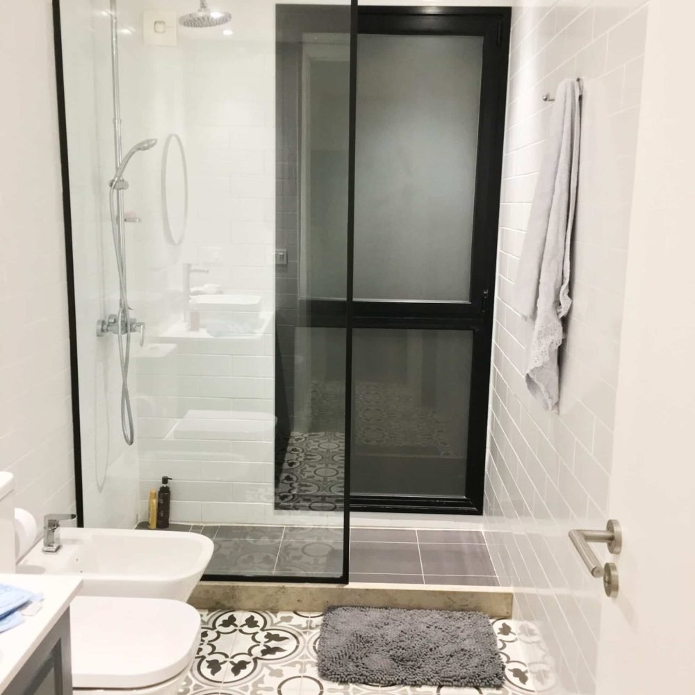 Baño contemporáneo en La Tahona, Canelones, Uruguay. Tonos grises y cálida iluminación, pisos de baldosa hidráulica. Mueble en silestone blanco y madera laqueada gris piedra. Espejo redondo con marco negro y mampara de ducha de vidrio amplio con perfilería en negro, destacando por su estilo moderno y funcionalidad.