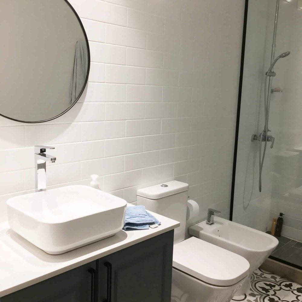 Baño contemporáneo en La Tahona, Canelones, Uruguay. Tonos grises y cálida iluminación, pisos de baldosa hidráulica. Mueble en silestone blanco y madera laqueada gris piedra. Espejo redondo con marco negro y mampara de ducha de vidrio amplio con perfilería en negro, destacando por su estilo moderno y funcionalidad.