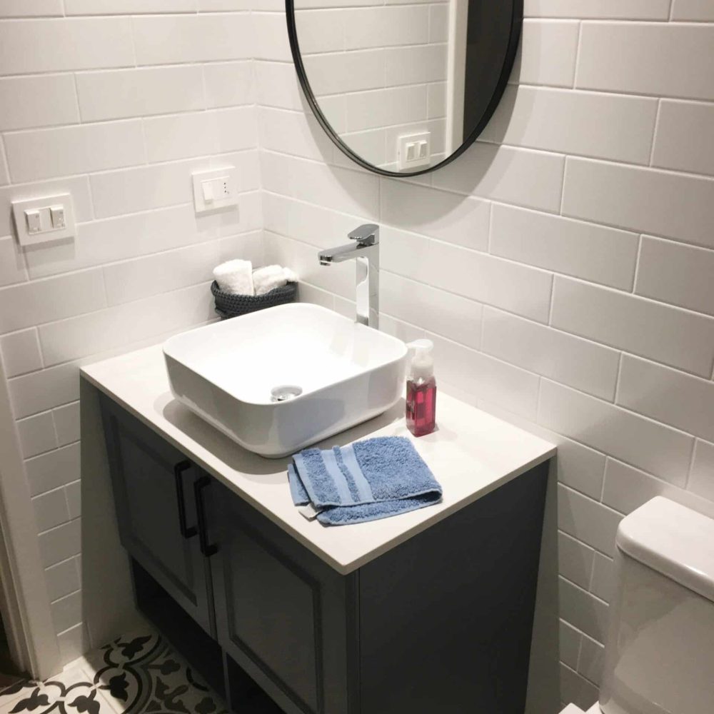 Baño contemporáneo en La Tahona, Canelones, Uruguay. Tonos grises y cálida iluminación, pisos de baldosa hidráulica. Mueble en silestone blanco y madera laqueada gris piedra. Espejo redondo con marco negro y mampara de ducha de vidrio amplio con perfilería en negro, destacando por su estilo moderno y funcionalidad.