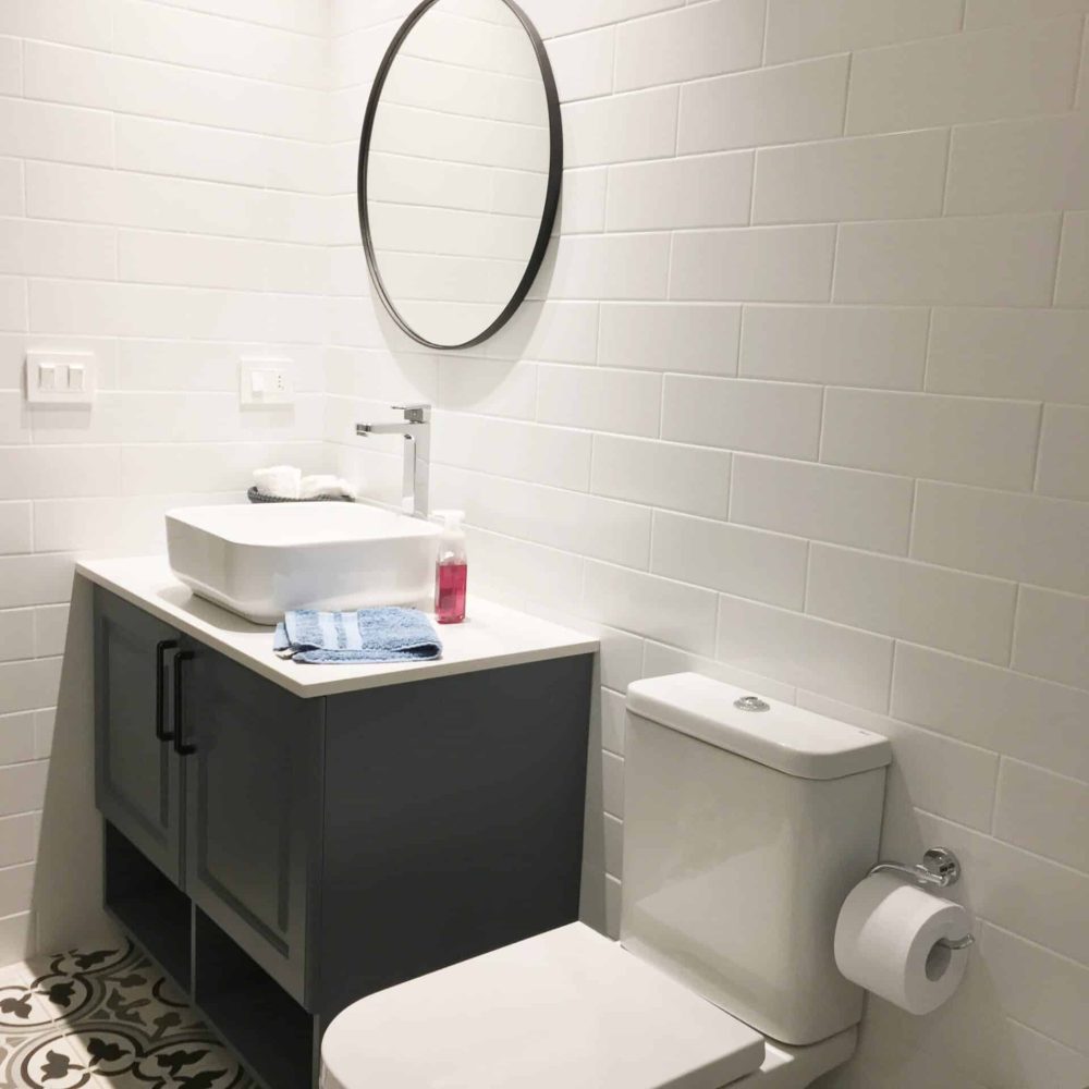 Baño contemporáneo en La Tahona, Canelones, Uruguay. Tonos grises y cálida iluminación, pisos de baldosa hidráulica. Mueble en silestone blanco y madera laqueada gris piedra. Espejo redondo con marco negro y mampara de ducha de vidrio amplio con perfilería en negro, destacando por su estilo moderno y funcionalidad.