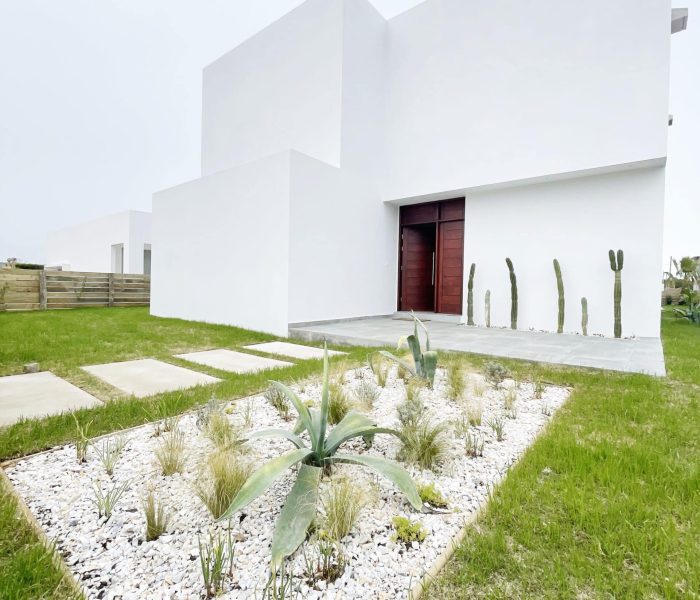 Casa La Mexicana Descubre la elegancia relajada de nuestra sala de estar en esta casa introspectiva con vista a la piscina en la zona de playas de José Ignacio. Este espacio, que incorpora un comedor, se caracteriza por su diseño funcional y acogedor. Un mueble multifuncional de 5 metros sirve como aparador para el comedor y como escritorio, albergando incluso una enfriadora de vinos para añadir un toque de sofisticación. El centro de atención de la sala es un cómodo sofá chaise en forma de L con una funda de tussor, acompañado de una butaca complementaria para mayor comodidad. Las cortinas de lino suavizan la luz natural, creando un ambiente sereno y acogedor perfecto para relajarse y disfrutar de la vista al aire libre.