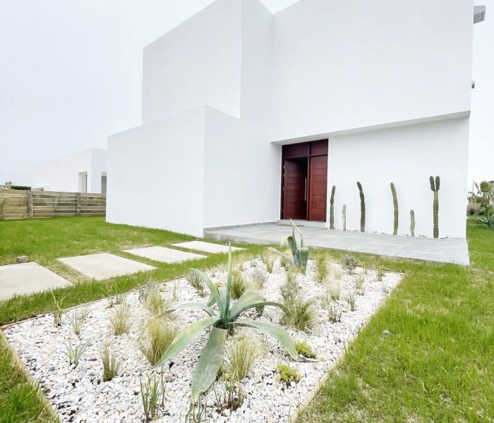 Descubre la elegancia relajada de nuestra sala de estar en esta casa introspectiva con vista a la piscina en la zona de playas de José Ignacio. Este espacio, que incorpora un comedor, se caracteriza por su diseño funcional y acogedor. Un mueble multifuncional de 5 metros sirve como aparador para el comedor y como escritorio, albergando incluso una enfriadora de vinos para añadir un toque de sofisticación. El centro de atención de la sala es un cómodo sofá chaise en forma de L con una funda de tussor, acompañado de una butaca complementaria para mayor comodidad. Las cortinas de lino suavizan la luz natural, creando un ambiente sereno y acogedor perfecto para relajarse y disfrutar de la vista al aire libre.