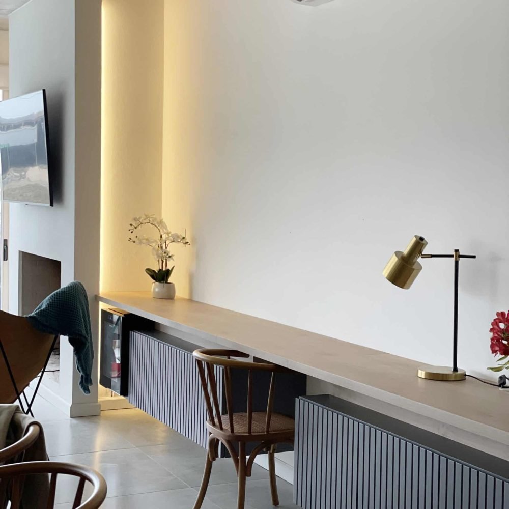 Descubre la elegancia relajada de nuestra sala de estar en esta casa introspectiva con vista a la piscina en la zona de playas de José Ignacio. Este espacio, que incorpora un comedor, se caracteriza por su diseño funcional y acogedor. Un mueble multifuncional de 5 metros sirve como aparador para el comedor y como escritorio, albergando incluso una enfriadora de vinos para añadir un toque de sofisticación. El centro de atención de la sala es un cómodo sofá chaise en forma de L con una funda de tussor, acompañado de una butaca complementaria para mayor comodidad. Las cortinas de lino suavizan la luz natural, creando un ambiente sereno y acogedor perfecto para relajarse y disfrutar de la vista al aire libre.