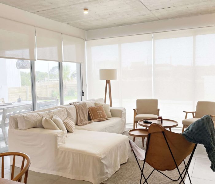 Sala de estar en José Ignacio Descubre la elegancia relajada de nuestra sala de estar en esta casa introspectiva con vista a la piscina en la zona de playas de José Ignacio. Este espacio, que incorpora un comedor, se caracteriza por su diseño funcional y acogedor. Un mueble multifuncional de 5 metros sirve como aparador para el comedor y como escritorio, albergando incluso una enfriadora de vinos para añadir un toque de sofisticación. El centro de atención de la sala es un cómodo sofá chaise en forma de L con una funda de tussor, acompañado de una butaca complementaria para mayor comodidad. Las cortinas de lino suavizan la luz natural, creando un ambiente sereno y acogedor perfecto para relajarse y disfrutar de la vista al aire libre.
