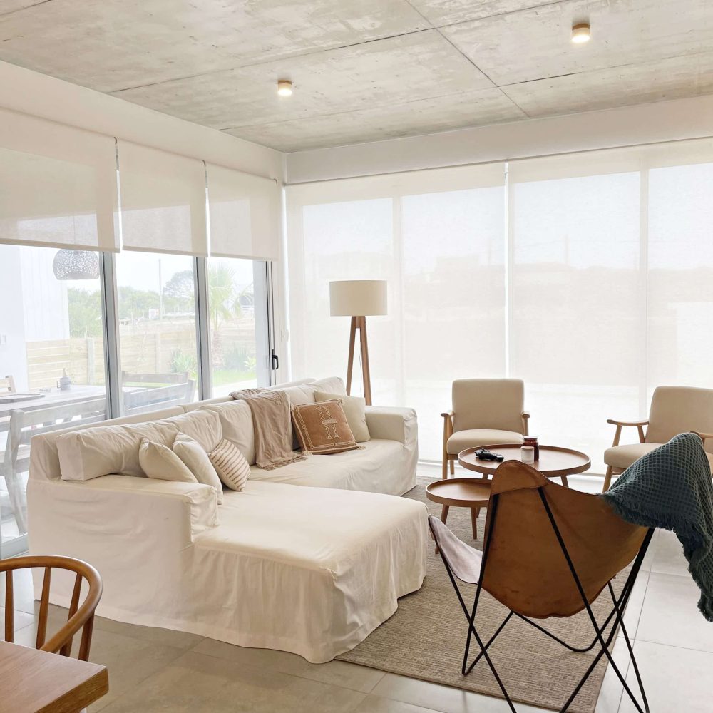Descubre la elegancia relajada de nuestra sala de estar en esta casa introspectiva con vista a la piscina en la zona de playas de José Ignacio. Este espacio, que incorpora un comedor, se caracteriza por su diseño funcional y acogedor. Un mueble multifuncional de 5 metros sirve como aparador para el comedor y como escritorio, albergando incluso una enfriadora de vinos para añadir un toque de sofisticación. El centro de atención de la sala es un cómodo sofá chaise en forma de L con una funda de tussor, acompañado de una butaca complementaria para mayor comodidad. Las cortinas de lino suavizan la luz natural, creando un ambiente sereno y acogedor perfecto para relajarse y disfrutar de la vista al aire libre.
