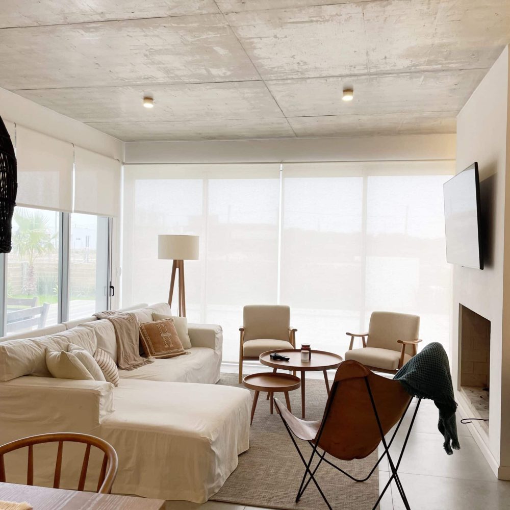 Descubre la elegancia relajada de nuestra sala de estar en esta casa introspectiva con vista a la piscina en la zona de playas de José Ignacio. Este espacio, que incorpora un comedor, se caracteriza por su diseño funcional y acogedor. Un mueble multifuncional de 5 metros sirve como aparador para el comedor y como escritorio, albergando incluso una enfriadora de vinos para añadir un toque de sofisticación. El centro de atención de la sala es un cómodo sofá chaise en forma de L con una funda de tussor, acompañado de una butaca complementaria para mayor comodidad. Las cortinas de lino suavizan la luz natural, creando un ambiente sereno y acogedor perfecto para relajarse y disfrutar de la vista al aire libre.