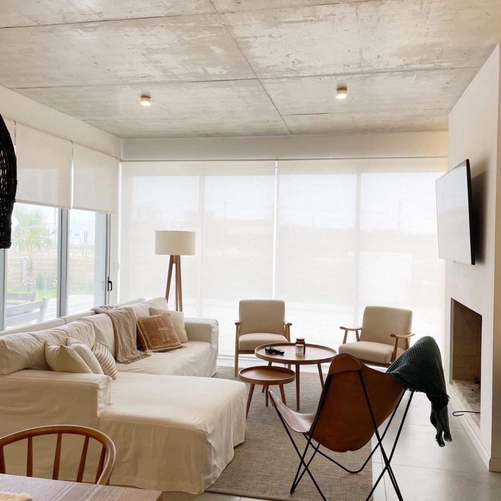 Descubre la elegancia relajada de nuestra sala de estar en esta casa introspectiva con vista a la piscina en la zona de playas de José Ignacio. Este espacio, que incorpora un comedor, se caracteriza por su diseño funcional y acogedor. Un mueble multifuncional de 5 metros sirve como aparador para el comedor y como escritorio, albergando incluso una enfriadora de vinos para añadir un toque de sofisticación. El centro de atención de la sala es un cómodo sofá chaise en forma de L con una funda de tussor, acompañado de una butaca complementaria para mayor comodidad. Las cortinas de lino suavizan la luz natural, creando un ambiente sereno y acogedor perfecto para relajarse y disfrutar de la vista al aire libre.