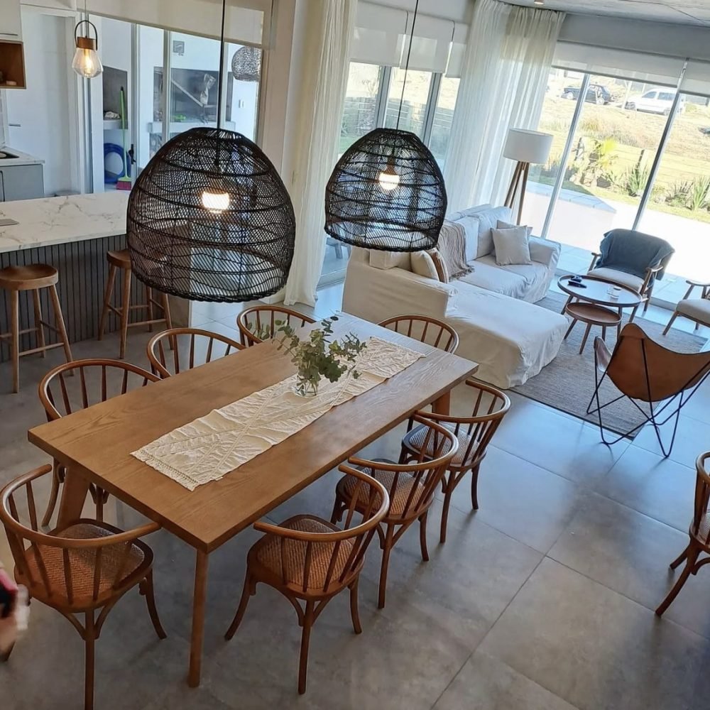 Descubre la elegancia relajada de nuestra sala de estar en esta casa introspectiva con vista a la piscina en la zona de playas de José Ignacio. Este espacio, que incorpora un comedor, se caracteriza por su diseño funcional y acogedor. Un mueble multifuncional de 5 metros sirve como aparador para el comedor y como escritorio, albergando incluso una enfriadora de vinos para añadir un toque de sofisticación. El centro de atención de la sala es un cómodo sofá chaise en forma de L con una funda de tussor, acompañado de una butaca complementaria para mayor comodidad. Las cortinas de lino suavizan la luz natural, creando un ambiente sereno y acogedor perfecto para relajarse y disfrutar de la vista al aire libre.