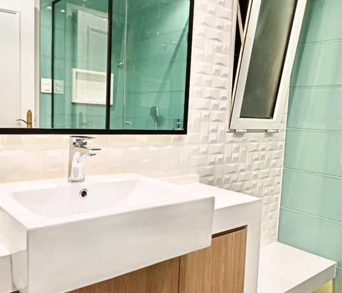 Baño contemporáneo en el barrio del Prado, Montevideo. Revestimientos en cerámica verde aguamarina, mesada de cuarzo blanco convertida en banco, mueble de roble claro, y cabina de ducha con cristal transparente y perfilería negra. Iluminación ambiental detrás del espejo y mueble flotante para crear un ambiente acogedor y moderno.