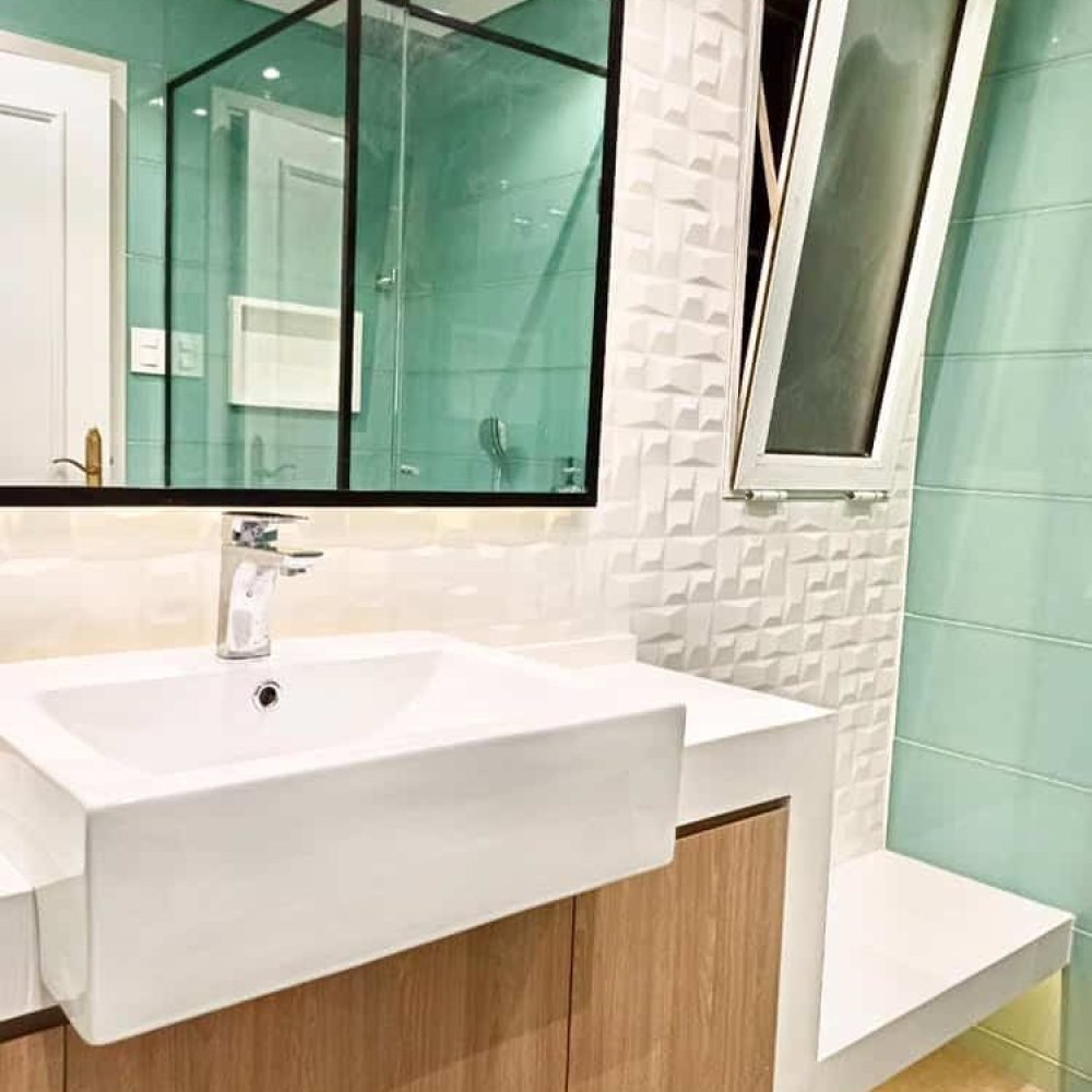 Baño contemporáneo en el barrio del Prado, Montevideo. Revestimientos en cerámica verde aguamarina, mesada de cuarzo blanco convertida en banco, mueble de roble claro, y cabina de ducha con cristal transparente y perfilería negra. Iluminación ambiental detrás del espejo y mueble flotante para crear un ambiente acogedor y moderno.